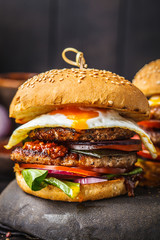 Homemade meat burger with egg, sauce and vegetables on dark background.