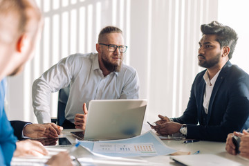 Smiling confident executive in white shirt sitting with laptop at meeting table with business partners team, discussing the working issues.