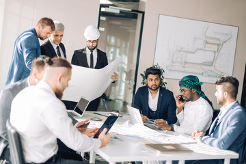 Fototapeta na wymiar Multiracial group of constructors, builders, engeneers and architects discussing blueprint at office. All men are dressed in business suits, and white shirts, one of them wears hardhat on head