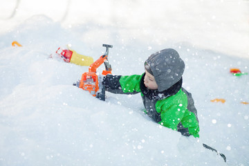 Portrait of cute little toddler sitting on snow and playing with his yellow tractor toy in the park. Child playing outdoors. Happy boy with construction toy. Lifestyle concept