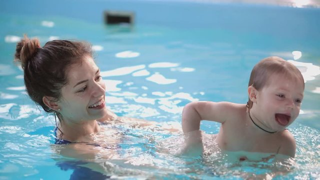 Cute little baby and his mother having swimming lesson in the pool. The Mother is holding his son in his hands and embracing him. Little boy is happily smiling
