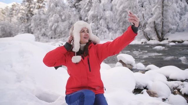 Beautiful girl in the mountains near winter river makes selfie on smartphone and smiling.