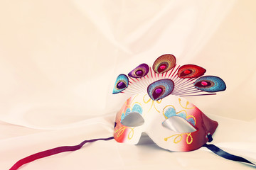 Photo of elegant and delicate venetian mask with peacock tail decoration element over white silk background. Vintage filtered.
