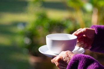 Close-up of hands elderly woman holding white coffee cup in the garden.