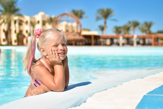 A girl in a bathing suit sits in the azure water of a swimming pool in a hotel with palm trees