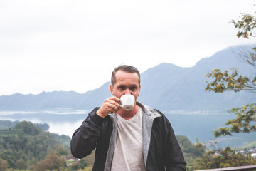 Young man traveler with white cup of coffee on a mountain background. Bali island.