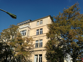 Schöner restaurierter Altbau in Beige und Naturfarben mit alten Bäumen einer Allee vor blauem...
