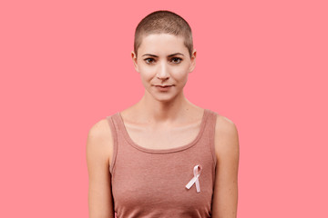 Smiling mid 30s woman, a cancer survivor, wearing pink breast cancer awareness ribbon, isolated...