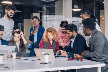 Creative multiracial business team discussing project, standing around workplace, while two co-workers with portable computers looking for needed information, sharing results to the workers