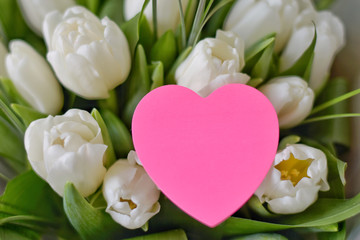 Bouquet of white beautiful tulips with tender petals and fresh green leaves with selective focus with blurred pink heart greeting card. Bunch of flower for romantic valentines or mothers day. 