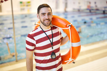 Portrait of smiling swim instructor standing and holding lifebuoy on his shoulder in the pool