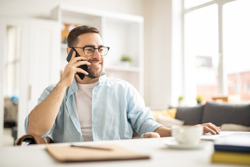 Happy young businessman with smartphone consulting with colleague while sitting by desk in front of...
