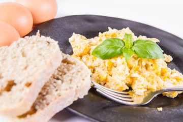 Scrambled eggs, some fresh eggs and wholemeal bread, and a fork, decorated with basil, on a white background 