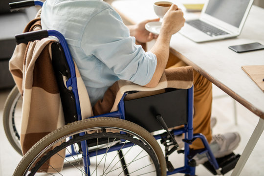 Businessman on wheelchair sitting by desk and having tea or coffee in front of laptop