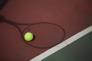 Tennis ball on a red-clay tennis court, lying in the shadow of tennis racket, being placed over it.