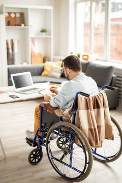 Businessman in casualwear making working notes in notepad while sitting in wheelchair at home