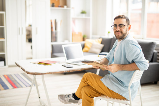 Happy young casual businessman with notepad sitting on chair by desk and working over new business project