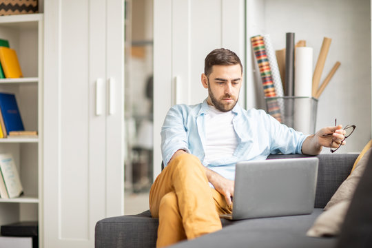 Young confident businessman in casualwear sitting on sofa in front of laptop at home and searching for online information
