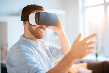 Happy young man with vr headset touching virtual stuff or pushing button on display while sitting...