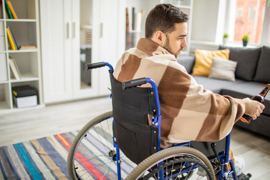 Sad young lonely man with bottle of beer or wine sitting in wheelchair in living-room