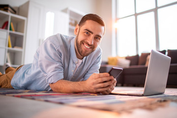 Cheerful young man in casualwear lying on the floor of living-room in front of laptop and scrolling in smartphone