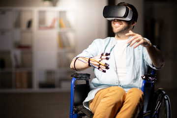 Young cheerful handicapped man with sensors and vr headset enjoying virtual adventure while sitting...