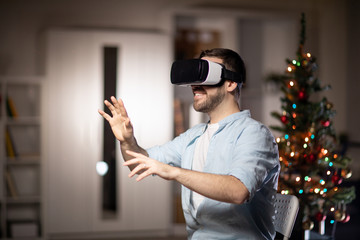 Cheerful young man with vr headset touching virtual stuff on display while sitting in dark room with sparkling xmas tree