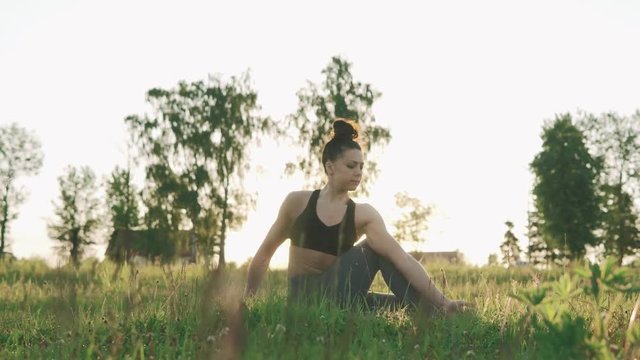 Brunette slender woman practicing yoga in morning park. Beautiful girl doing yoga moves on green grass near lake at sunrise with copy space