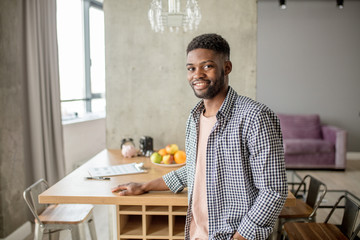 Happy young African bearded man in casual clothes looking at camera, posing over modern home interior.