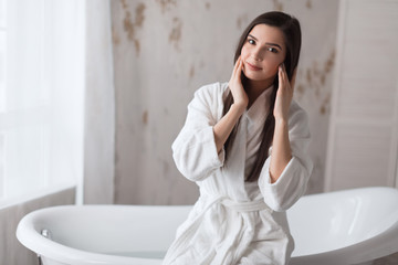 Brown-haired young woman in a bathrobe looking at camera and sitting on bathtub
