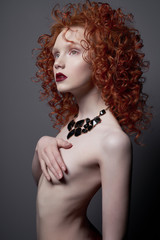 Beautiful young woman with black jewelry and red-hair on grey background. Nude pretty lady with bright red lipsstick and modern art makeup. Sensual model pose in photo-studio. Sexy naked body.