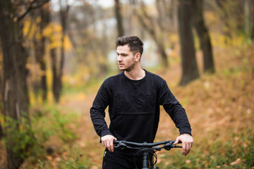 Young pretty athletic man standing with bicycle in colorful autumn park. Fall season background. Male cyclist on the road with fallen leaves