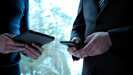 Two men (guys) are holding a tablet and a mobile in the hands of one of the men in a suit the...