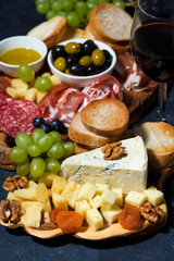 cheese platter on a wooden board, bread, fruit and cold cuts on dark background, vertical closeup