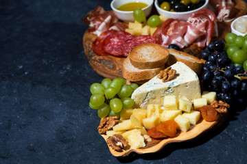 cheese platter on a wooden board, bread, fruit and cold cuts on dark background