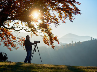 Male tourist photographer standing under large tree with golden leaves with tripod and professional camera to take picture of woody foggy mountains landscape and bright sunset in blue sky background.