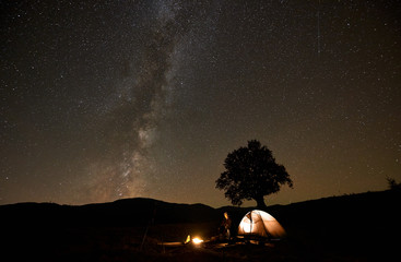 Summer night camping and astrophotography. Photographer man sitting in front of tourist tent and tripod camera at burning campfire watching beautiful dark starry sky with Milky Way constellation.
