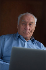 Senior grey-haired man using a laptop sitting in chair at home.