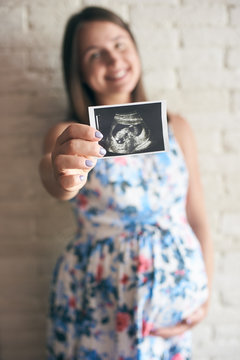 Selective focus of ultrasound image of baby in hand of smiling pregnant woman. Future parent standing, looking at camera and laughing at background. Concept of happiness and pregnancy.