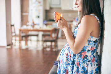 Side view of pregnant woman in dress standing in kitchen and keeping in hands cigarette and...
