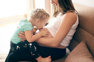 Side view of happy pregnant mother sitting on sofa and cute daughter kissing and embracing big belly. Lovely girl expecting little brother. Concept of parental love and happiness.
