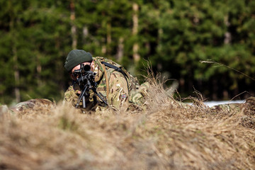 sniper laying on the ground covered in a ghille suite tall grass and trees in the background....