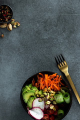 Vegan buddha bowl. Bowl with fresh raw vegetables - carrot, zucchini, tomatoes cherry and avocado, nuts. Dark grey background, top view, copy space
