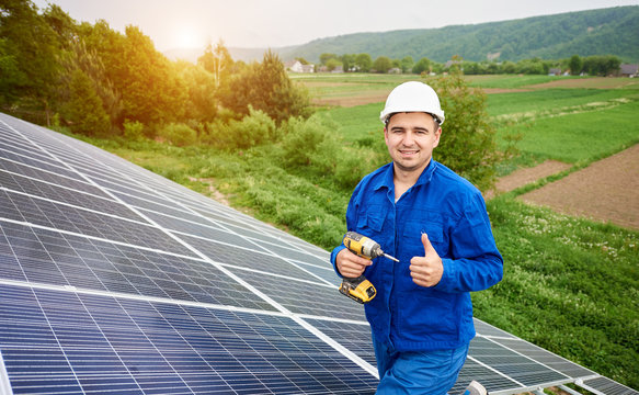 Close-up of construction worker with screwdriver with thumb-up gesture on photo voltaic panel solar system shiny surface and lit by sun green fields background. Alternative energy concept.