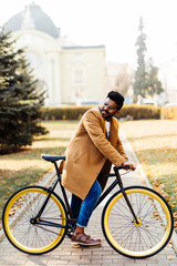 Fototapeta premium Side view of a young indian hipster man in coat walking with a bicycle on a city street