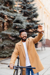 Young handsome indian hipster man in coat wave with hands hello gesture for greatings standing near his bicycle on street