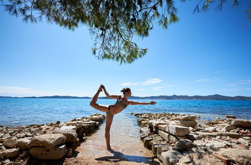 Young attractive suntanned woman in swimming suite making yoga pose near sea water by stony path under bright blue sky. Active lifestyle, summer holidays, recreation and vacations concept.