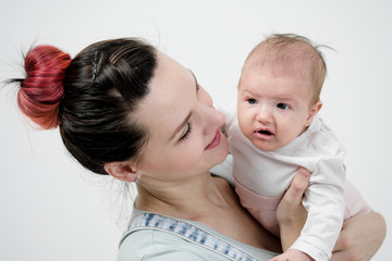 Young woman mother in denim overalls holds a baby child in her arms. White background in the studio.
