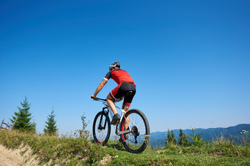Fototapeta na wymiar Back view of young athletic tourist biker in helmet and full equipment cycling bike up the grassy hill on distant mountains and blue summer sky background. Active lifestyle and extreme sport concept.