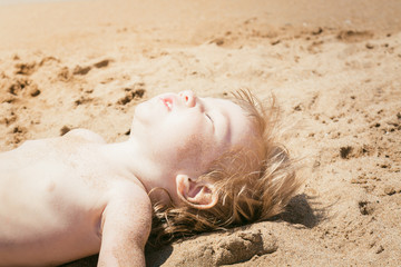 Small child sunbathes on hot sand with his eyes closed in sun.